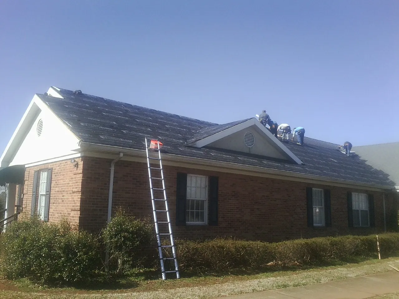 Crew tearing shingles off a brick ranch roof with a ladder set — Emergency Roof Repair work by our crew in Guilford